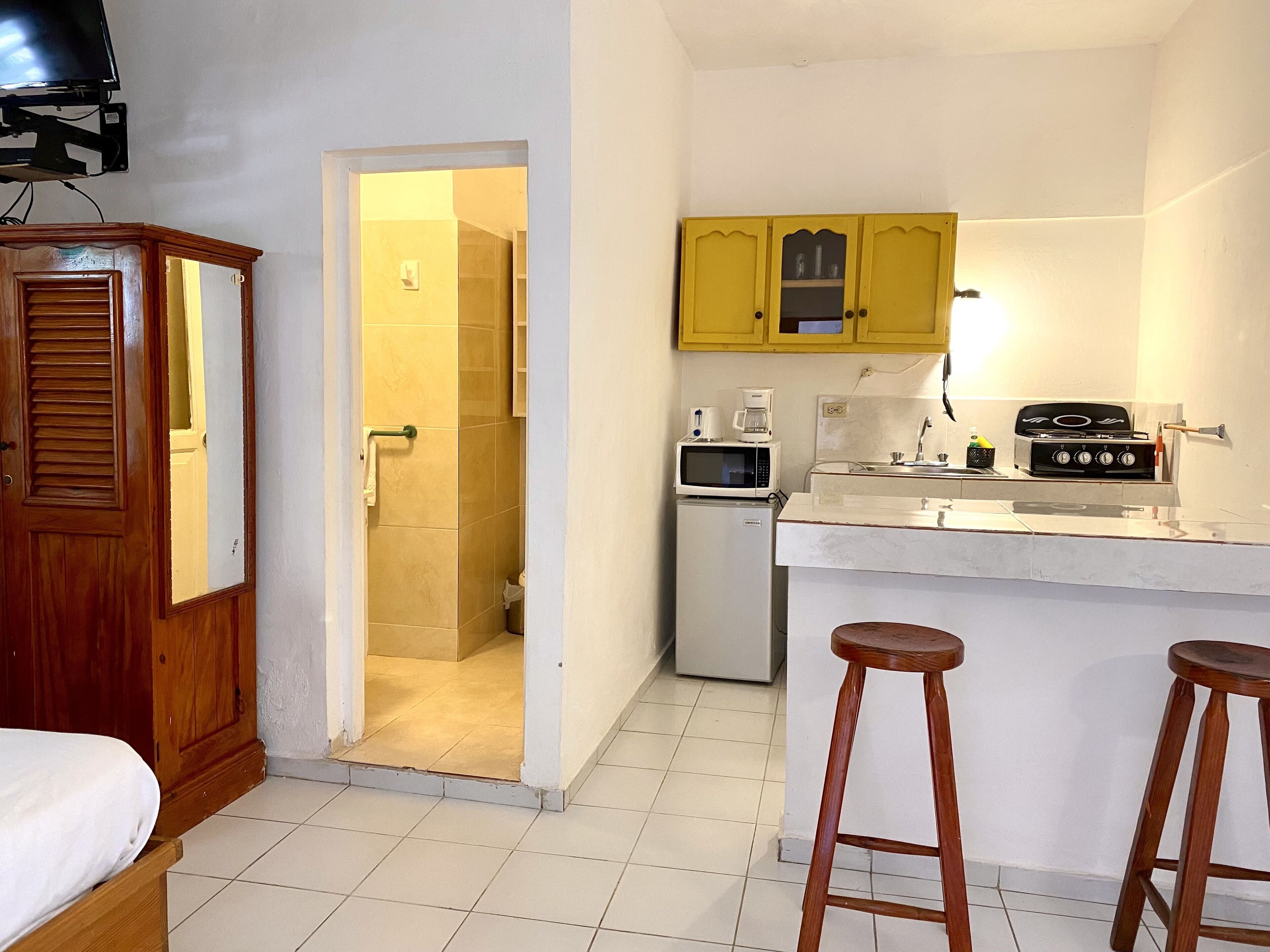 A compact kitchen with a refrigerator, microwave, stove, and yellow cabinets. A wooden door and a small stool are also visible.