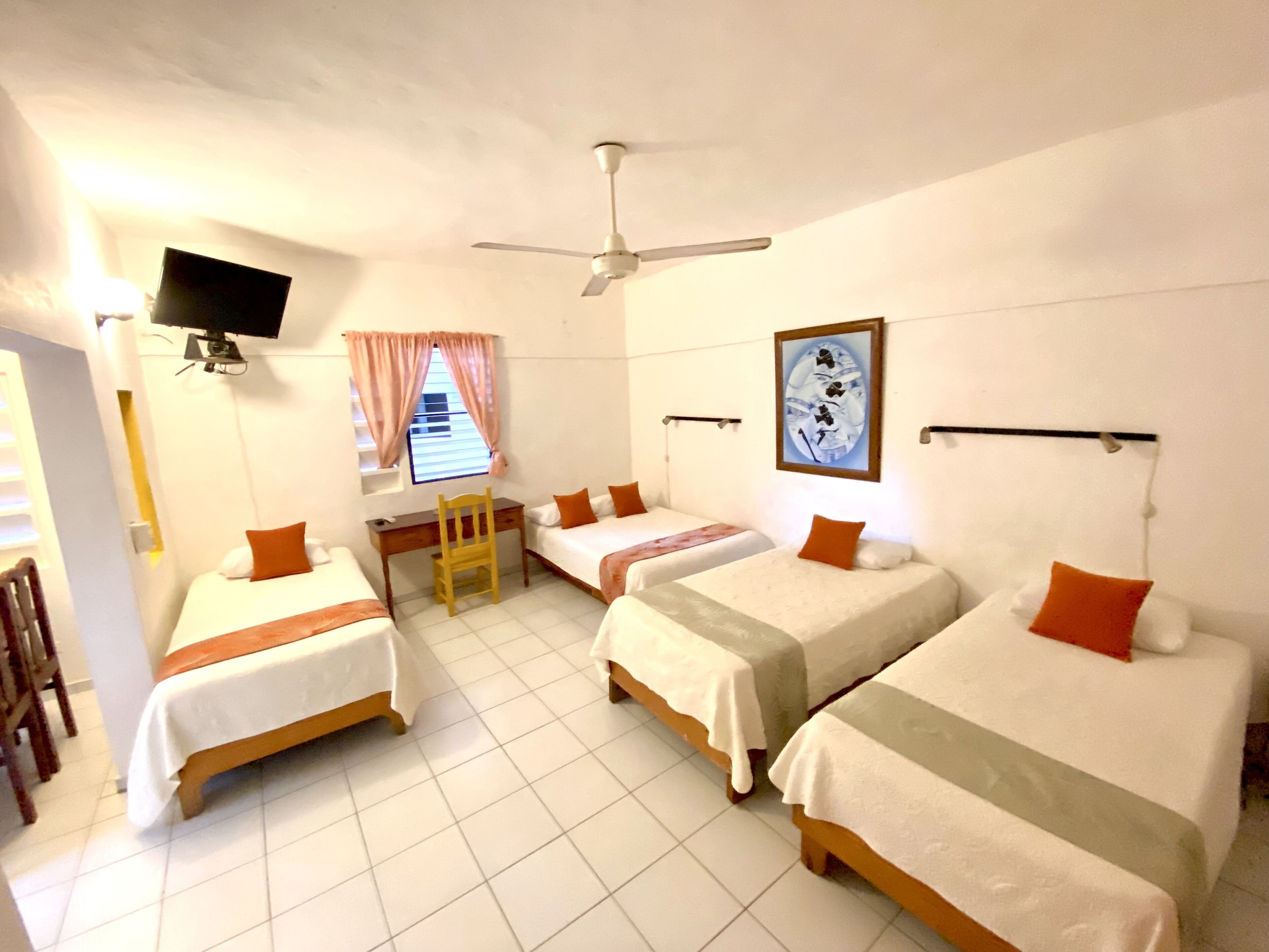 Habitación de hotel con tres camas individuales, un escritorio, una silla, un ventilador de techo, un televisor y una ventana con cortinas.