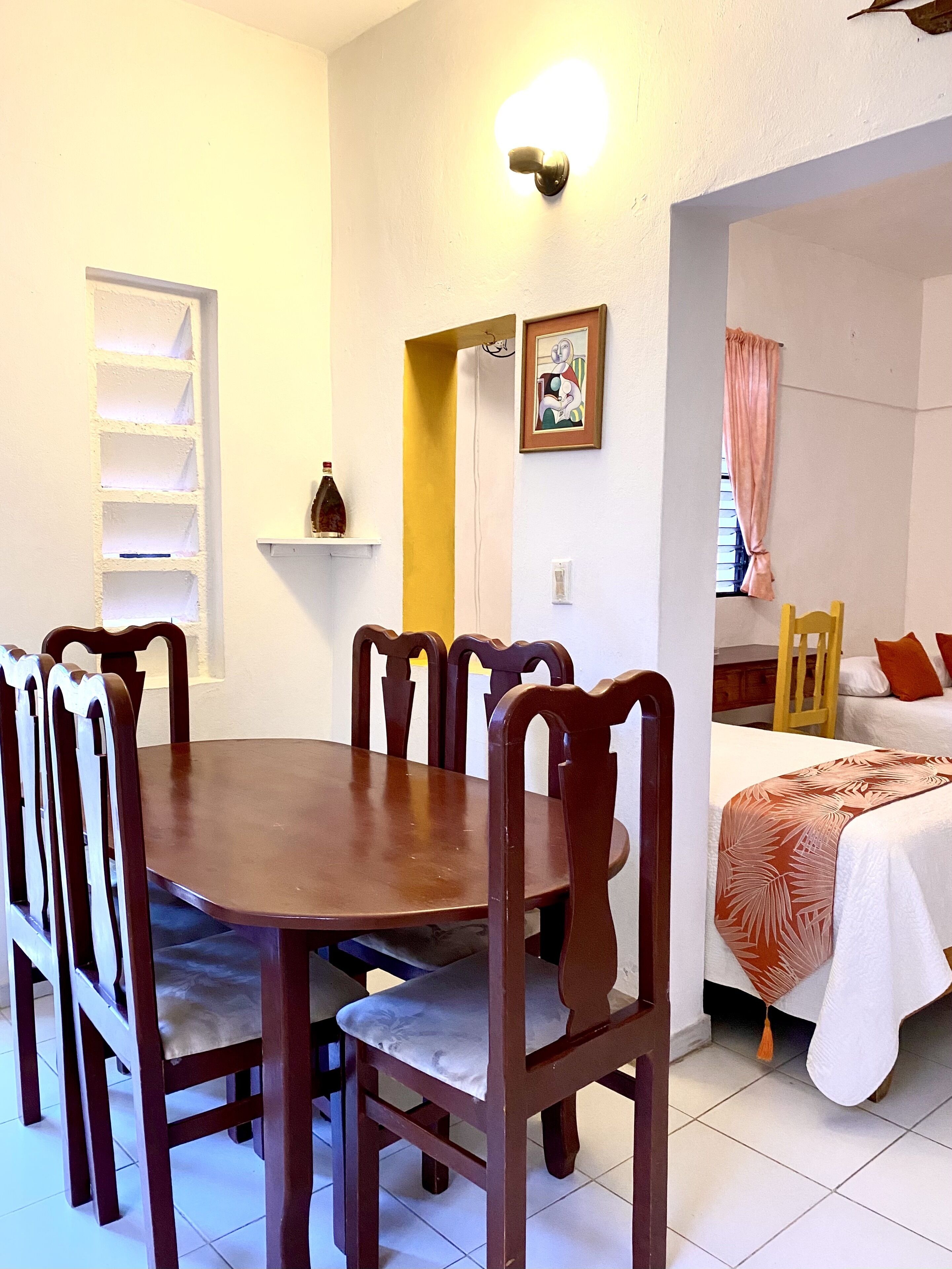 A dining area with a wooden table and chairs, a shelf with a bottle, a painting, and a bed with a patterned bedspread.