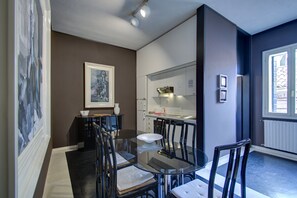 A modern dining area with a glass table and black chairs, a framed artwork on the wall, and a kitchen in the background.