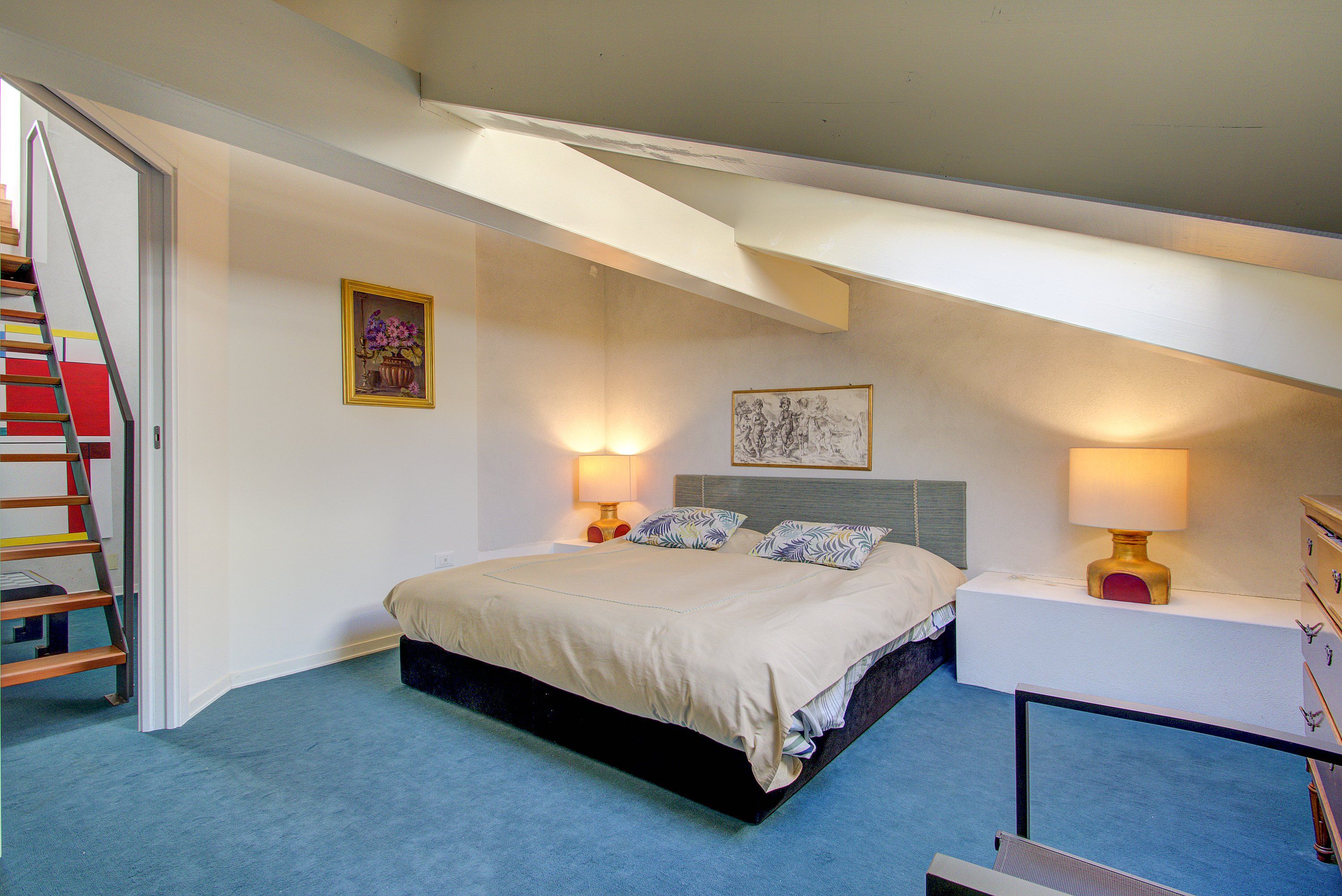 A bedroom with a bed, bedside lamps, a nightstand, a painting, and a staircase.
