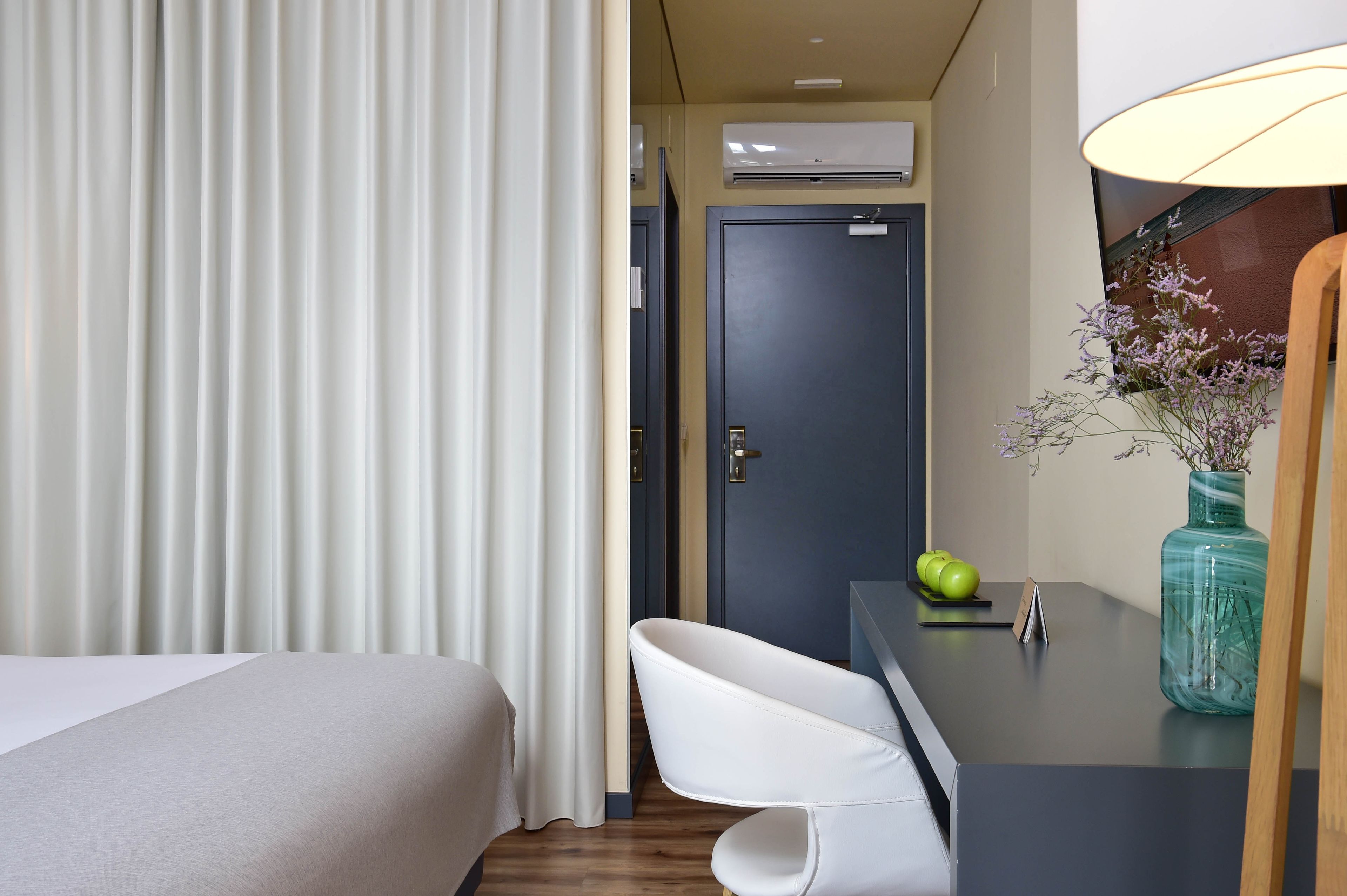 Quarto de hotel com uma cama, uma escrivaninha com luminária, um vaso com flores e uma porta.