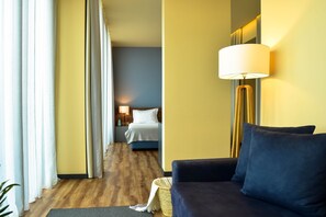 A hotel room with a dark blue sofa, a bed with white bedding, a lamp, and a wooden floor.