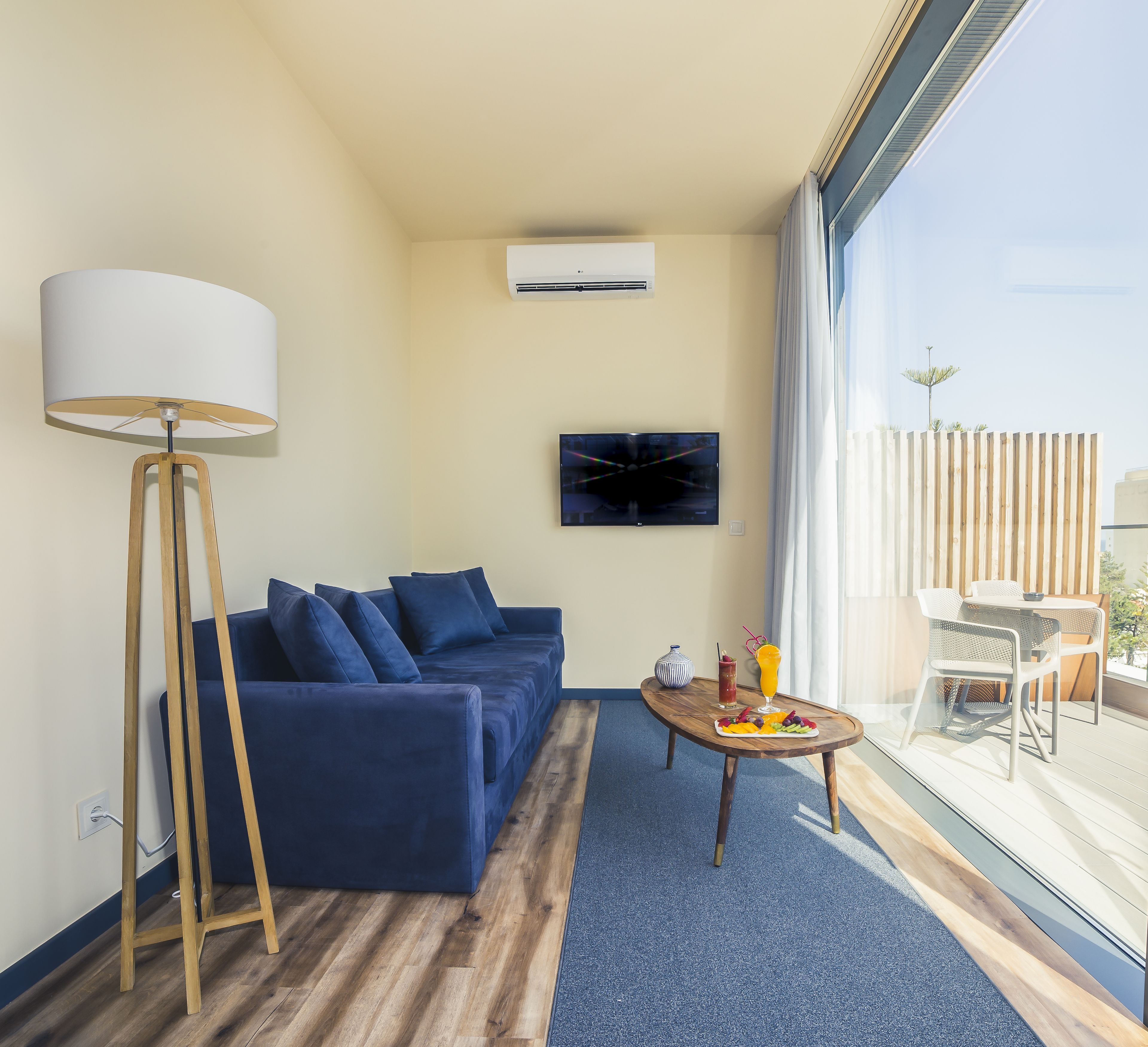 A modern living room with a blue sofa, a floor lamp, a flat-screen TV, and a dining area with a table and chairs.