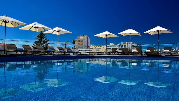 A poolside area with lounge chairs and umbrellas, a clear blue sky, and a building in the background. - The Prime Energize Hotel & Spa (Vila Real Santo Antonio)