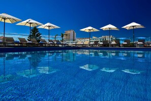 Un área junto a la piscina con sillas de descanso y sombrillas, un cielo azul despejado y un edificio al fondo.