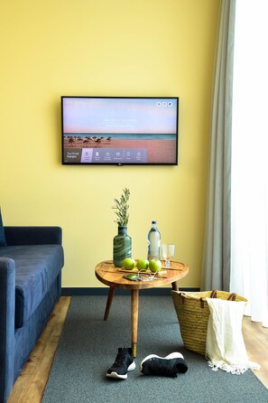 A room with a TV displaying a beach scene, a blue sofa, a small table with a vase, a bottle, and a glass, and a wicker basket.
