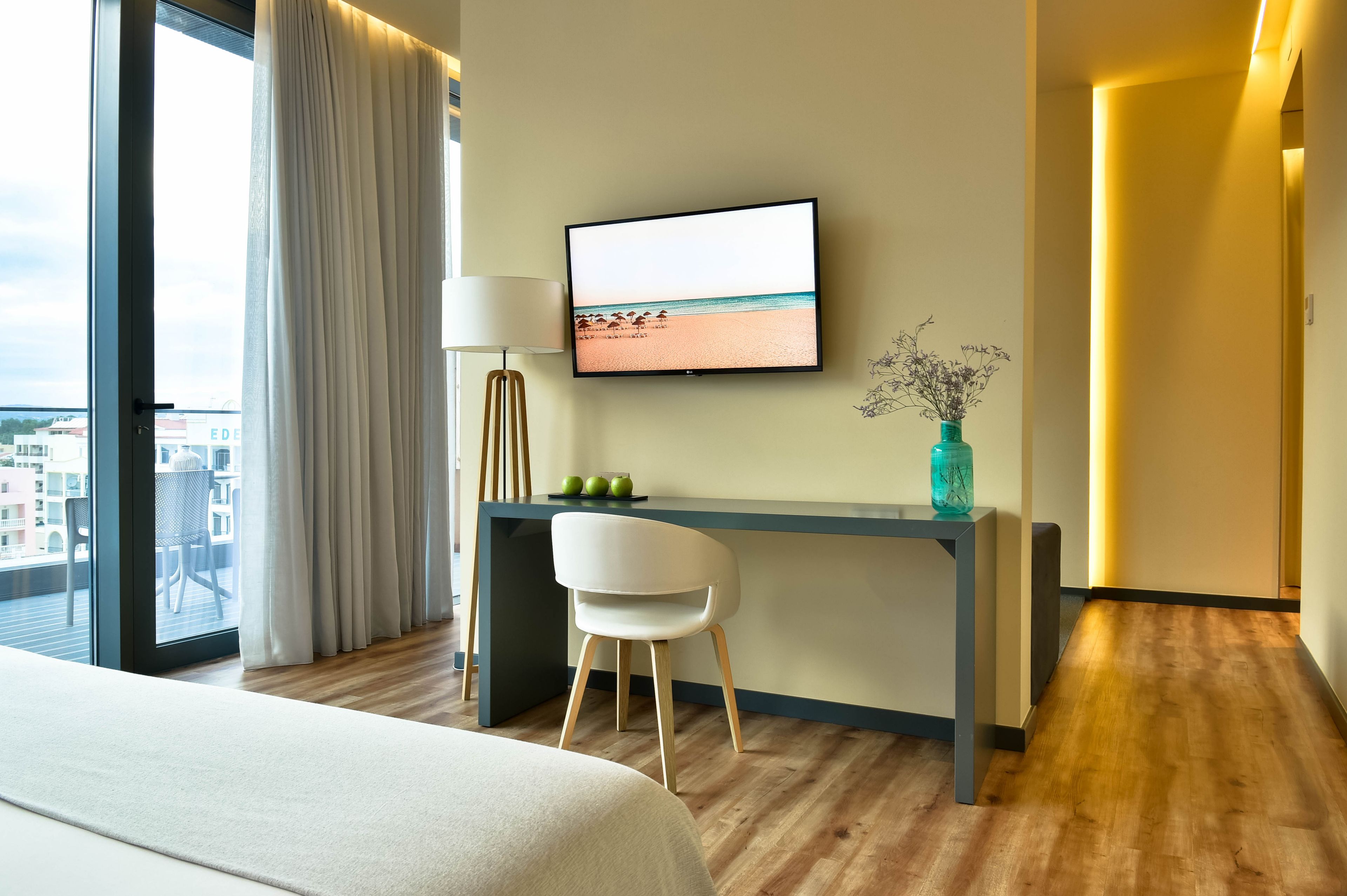 Une chambre d’hôtel avec un lit, un bureau équipé d’une télévision, une chaise, une lampe et une vue sur la ville grâce à une grande fenêtre.