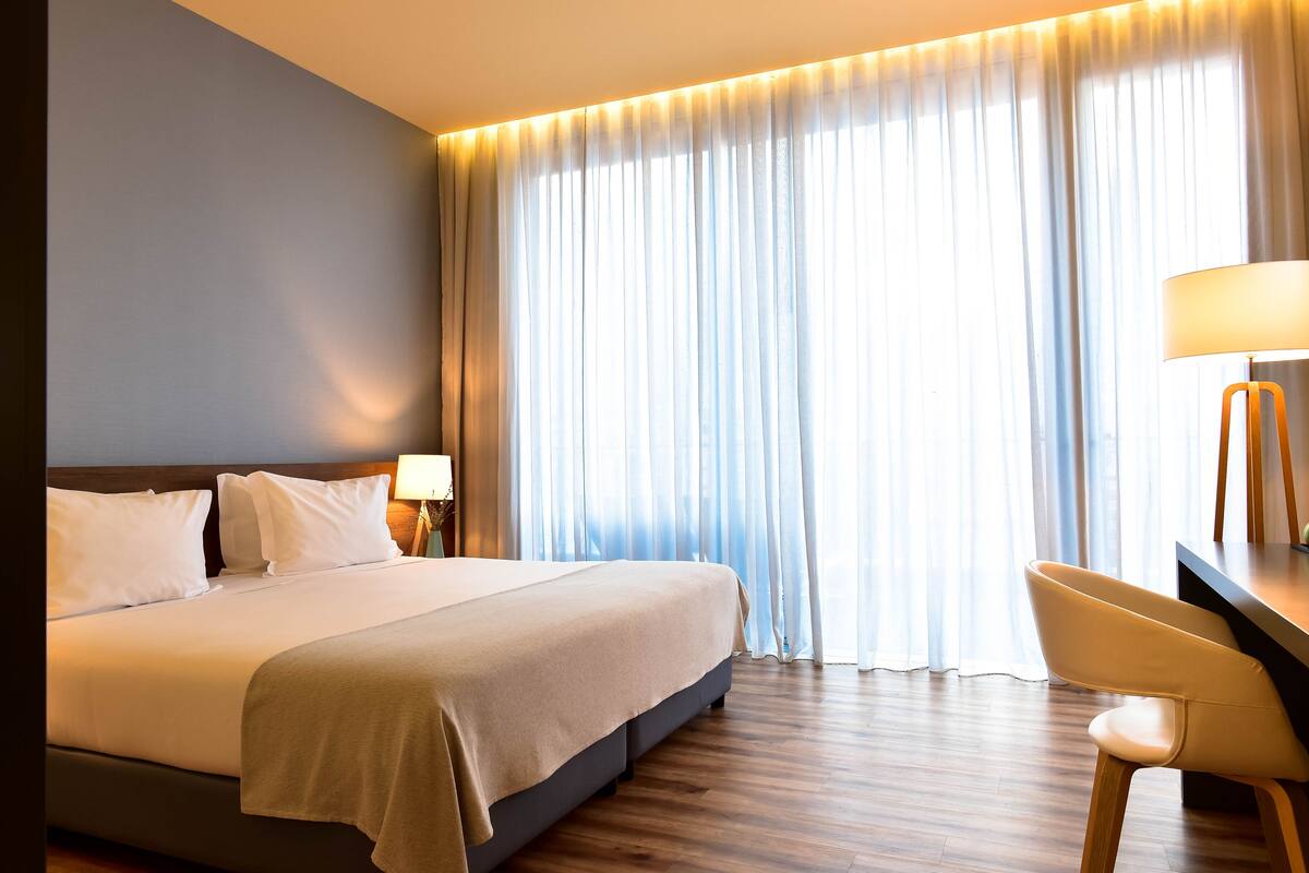 A hotel room with a large bed, a desk with a chair, a lamp, and a view through sheer curtains.