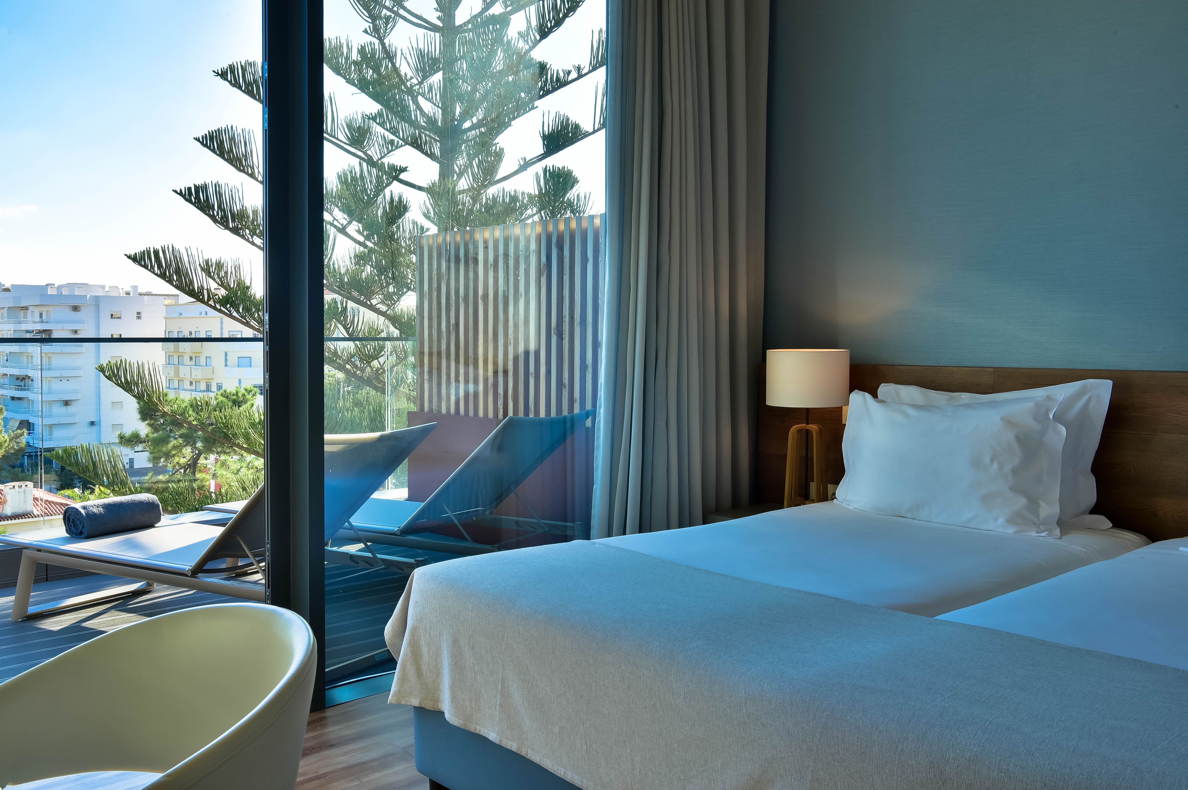A hotel room with a bed, a chair, a desk, and a view of buildings and trees outside the window.