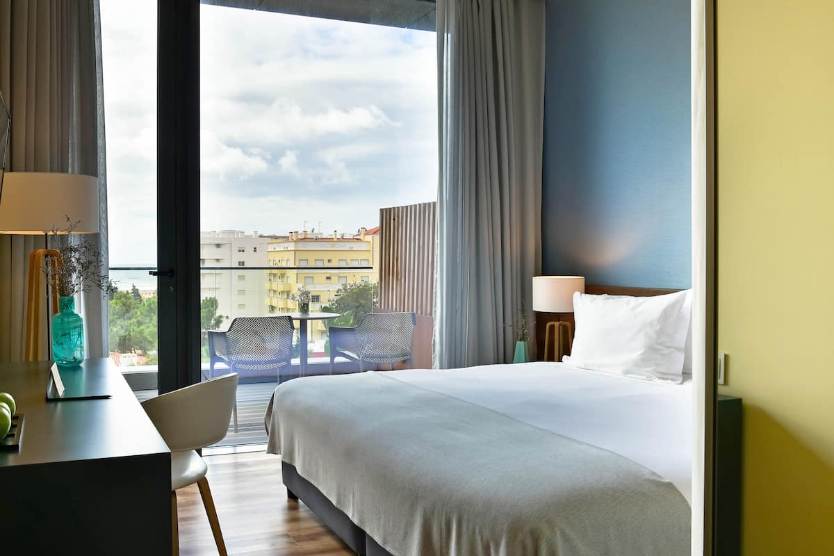 A hotel room with a large bed, a desk with a lamp, and a view of buildings through a window.