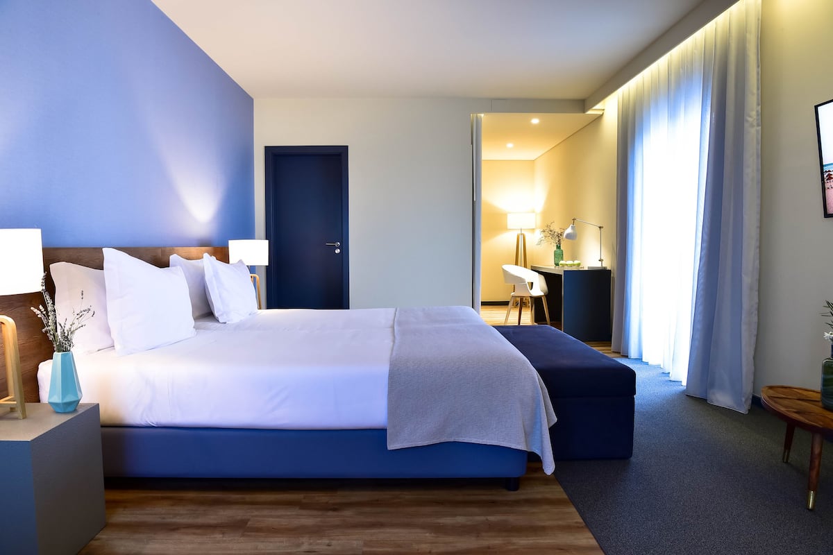 A modern hotel room with a large bed, bedside tables, a desk, and a view of the hallway.