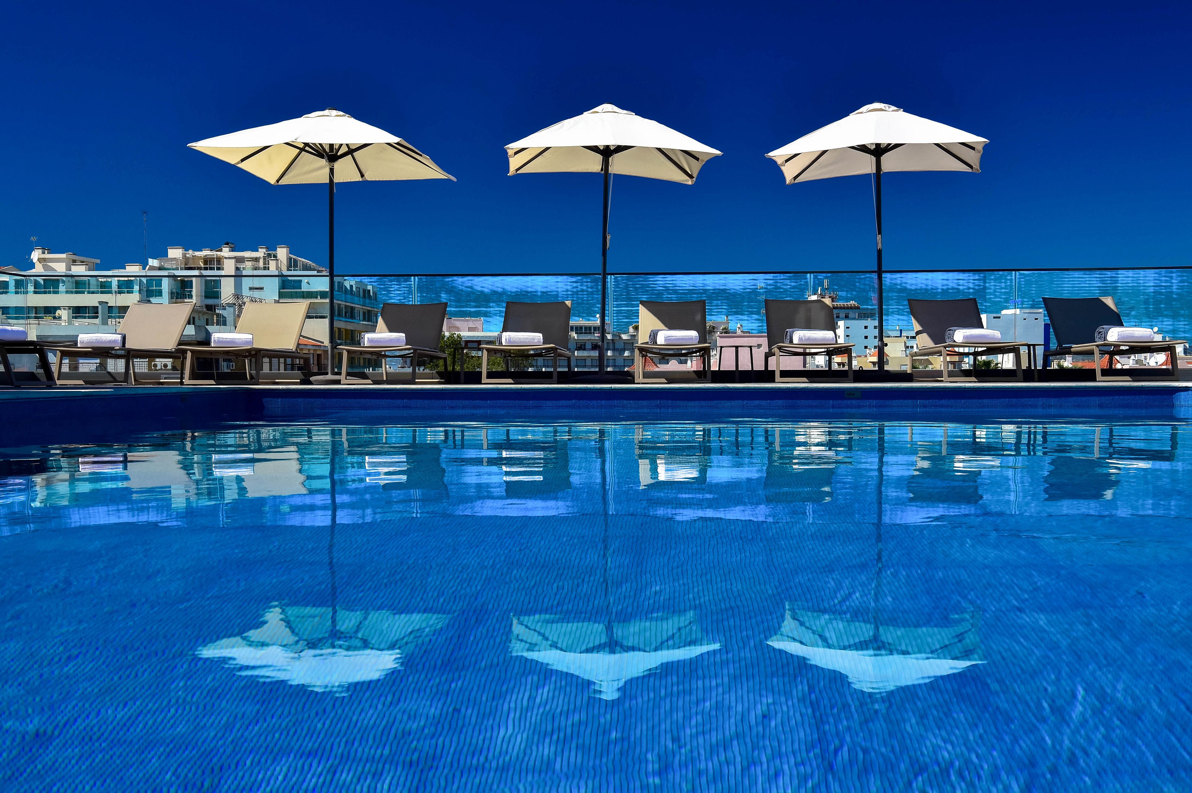 Zona de piscina con sillones y sombrillas, cielo azul despejado y un edificio al fondo.