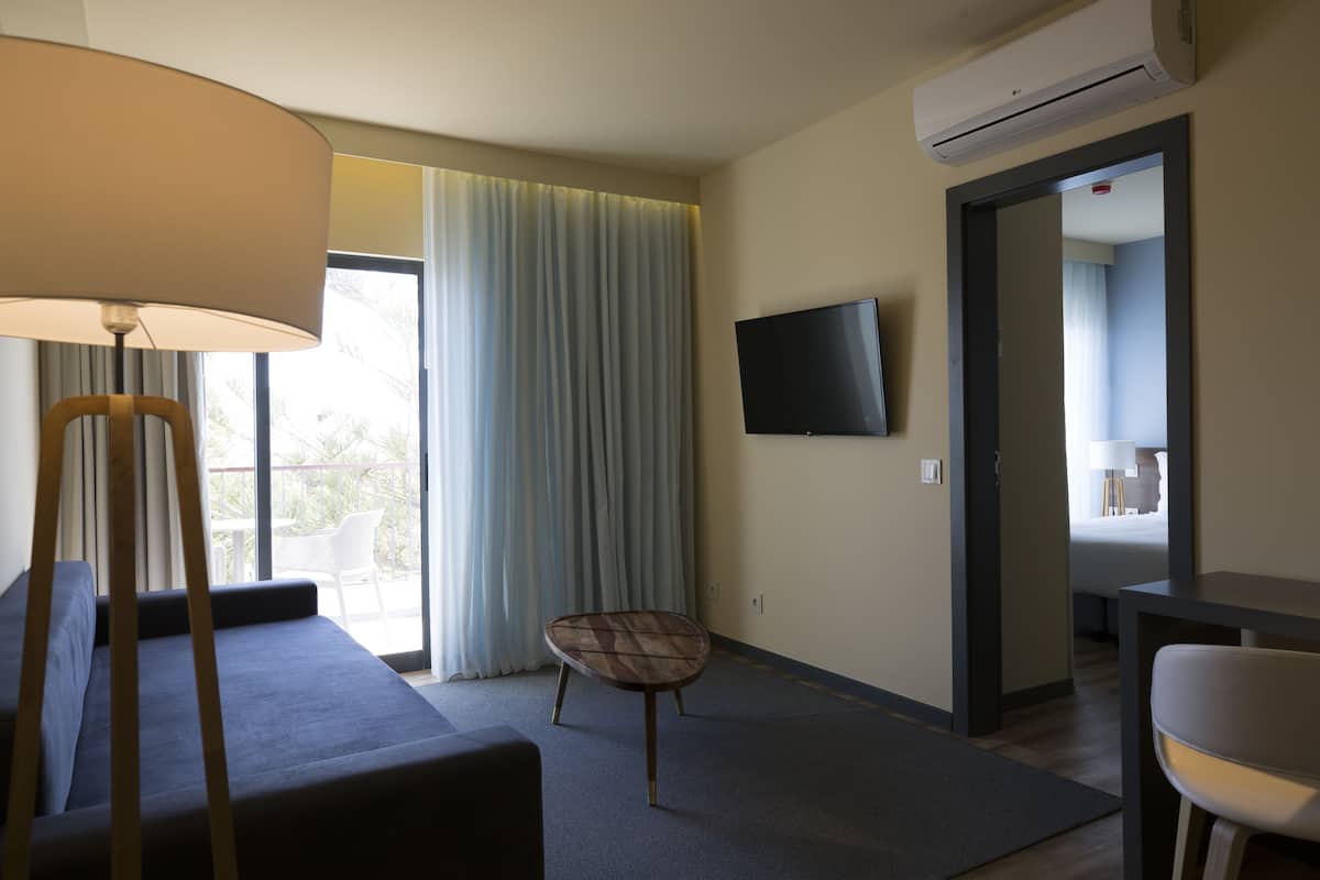 A hotel room with a sofa, a small table, a chair, a TV mounted on the wall, and a window with curtains.