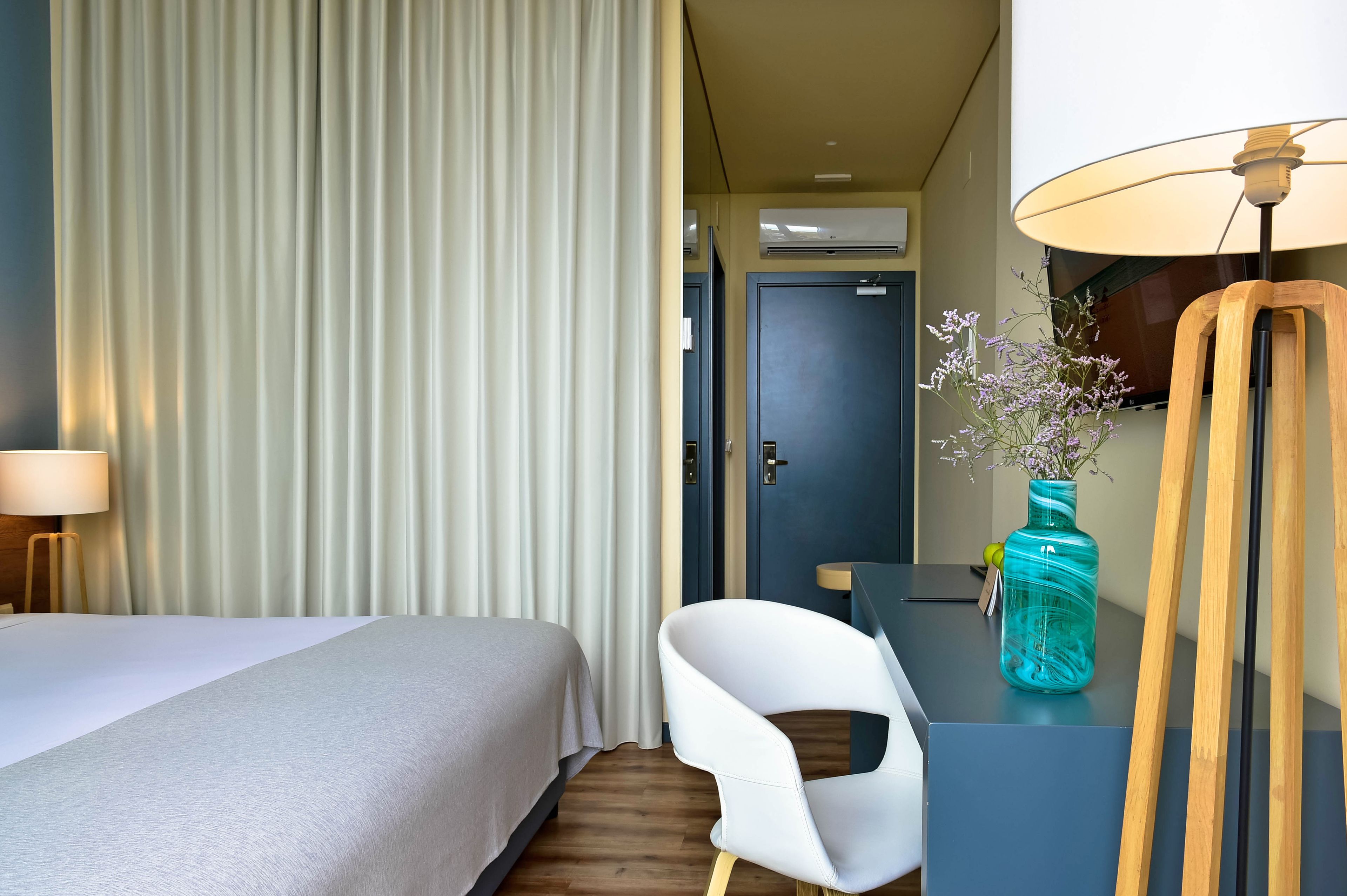 A hotel room with a bed, a white chair, a desk with a lamp, and a vase with flowers.