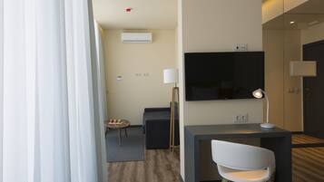 A modern hotel room with a flat-screen TV, a desk with a lamp, a chair, a small round table with fruits, and a sofa.