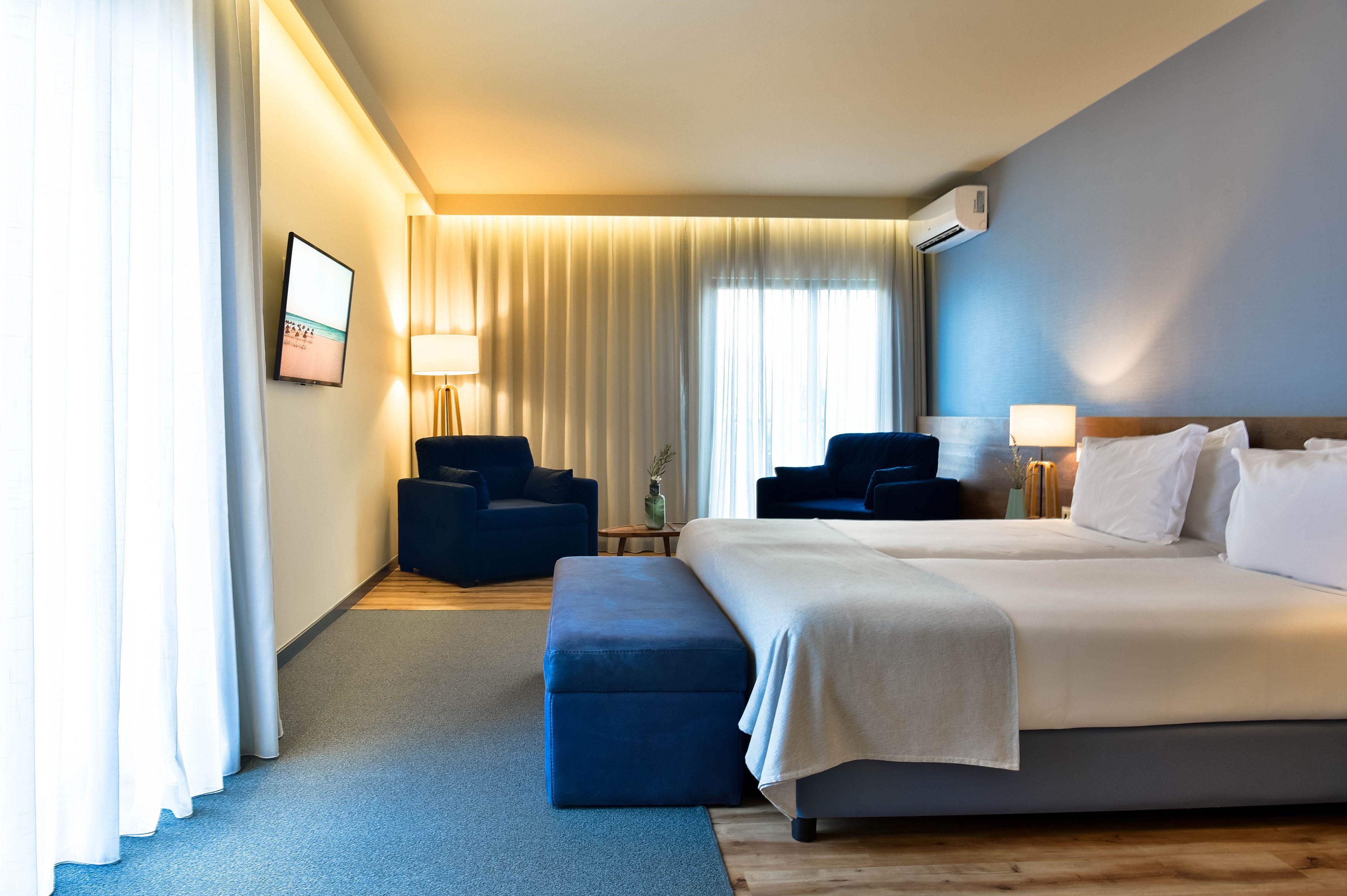 Ein Hotelzimmer mit einem großen Bett, zwei Sesseln, einem kleinen Tisch und einem Fernseher an der Wand.