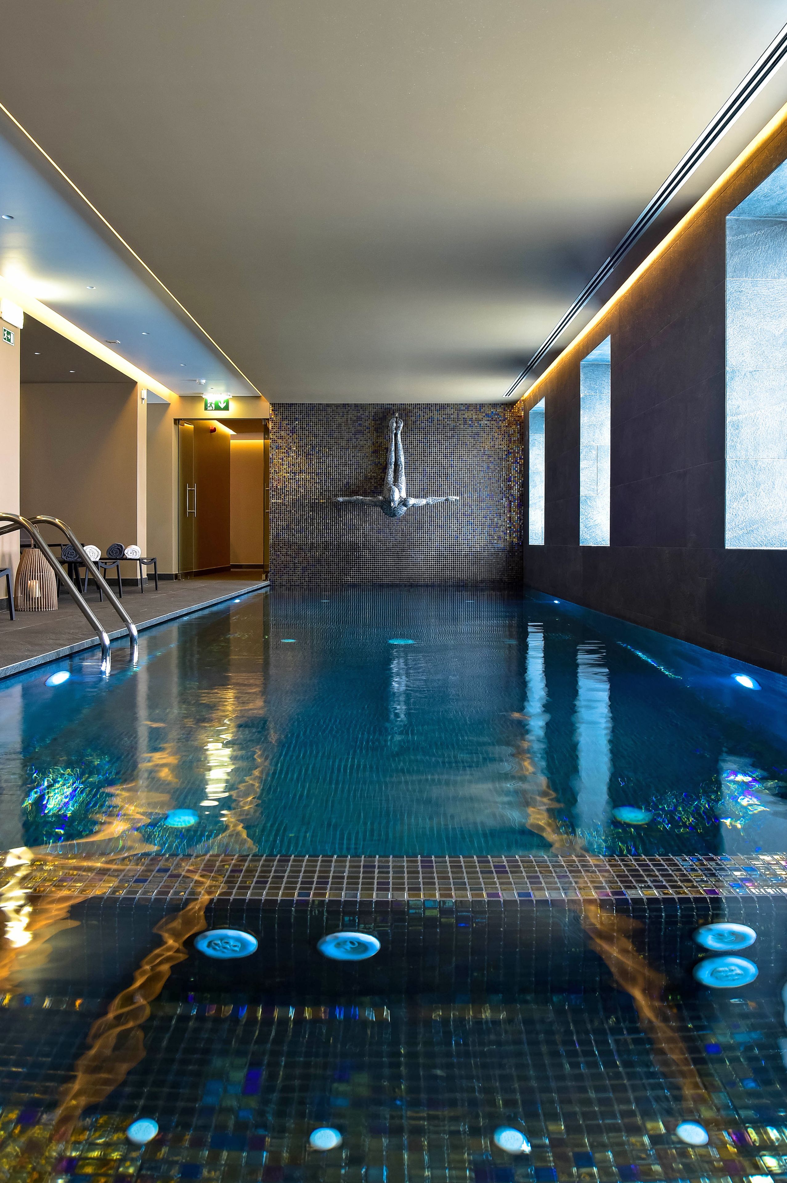 An indoor swimming pool with a tiled wall, a water feature, and a staircase leading to a door.