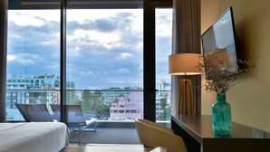 A hotel room with a bed, a desk with a lamp, a vase with flowers, and a view of the city through large windows. - The Prime Energize Hotel & Spa (Vila Real Santo Antonio)
