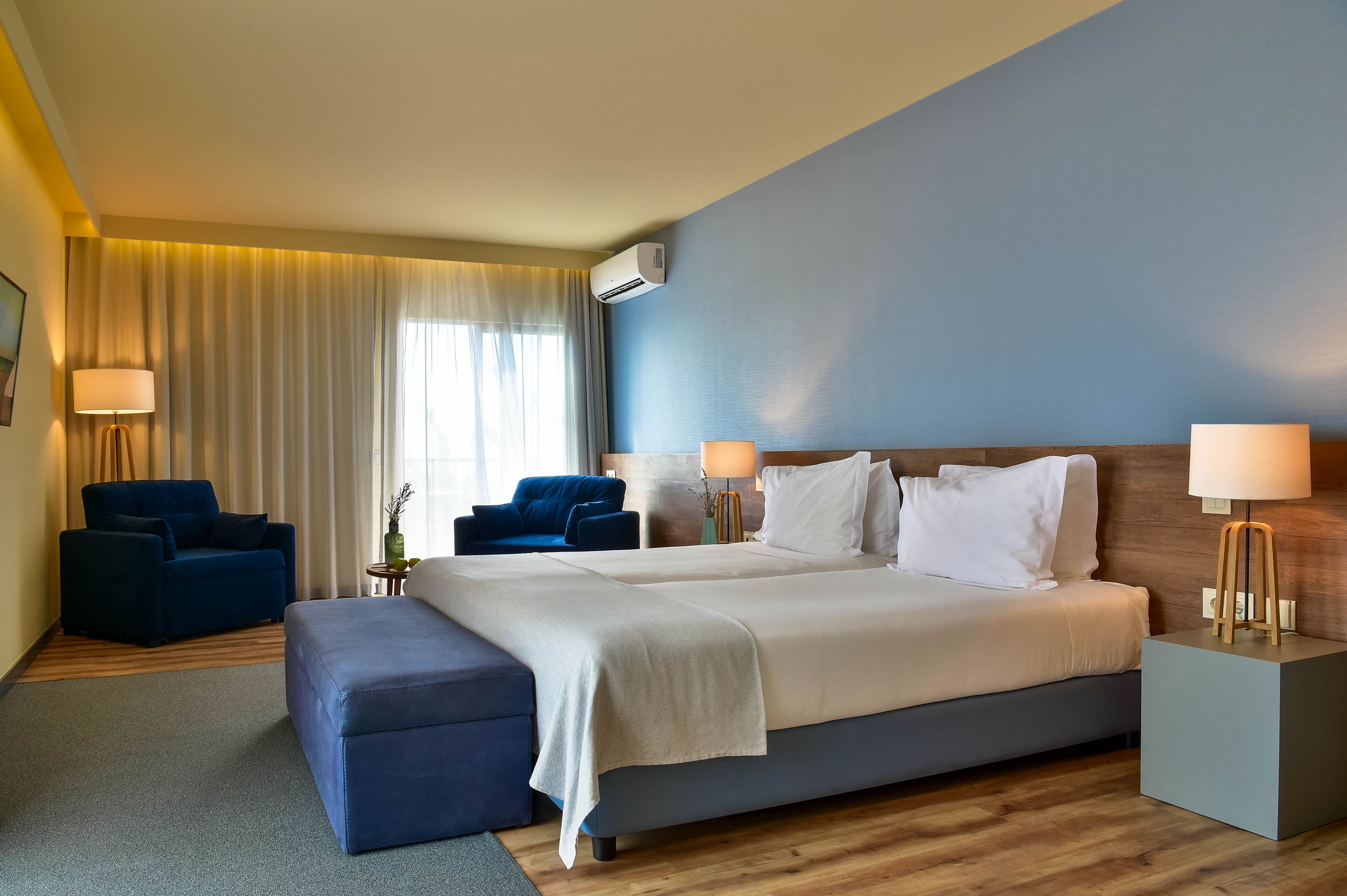 A modern hotel room with a large bed, two armchairs, a small table, and a bedside lamp.