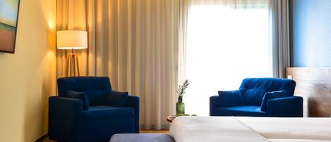 Una habitación de hotel con una cama, dos sillones azules, una lámpara de pie y un ventanal con cortinas.