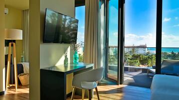 Habitación de hotel moderna con una televisión grande, un escritorio, una silla y vistas al océano y edificios.