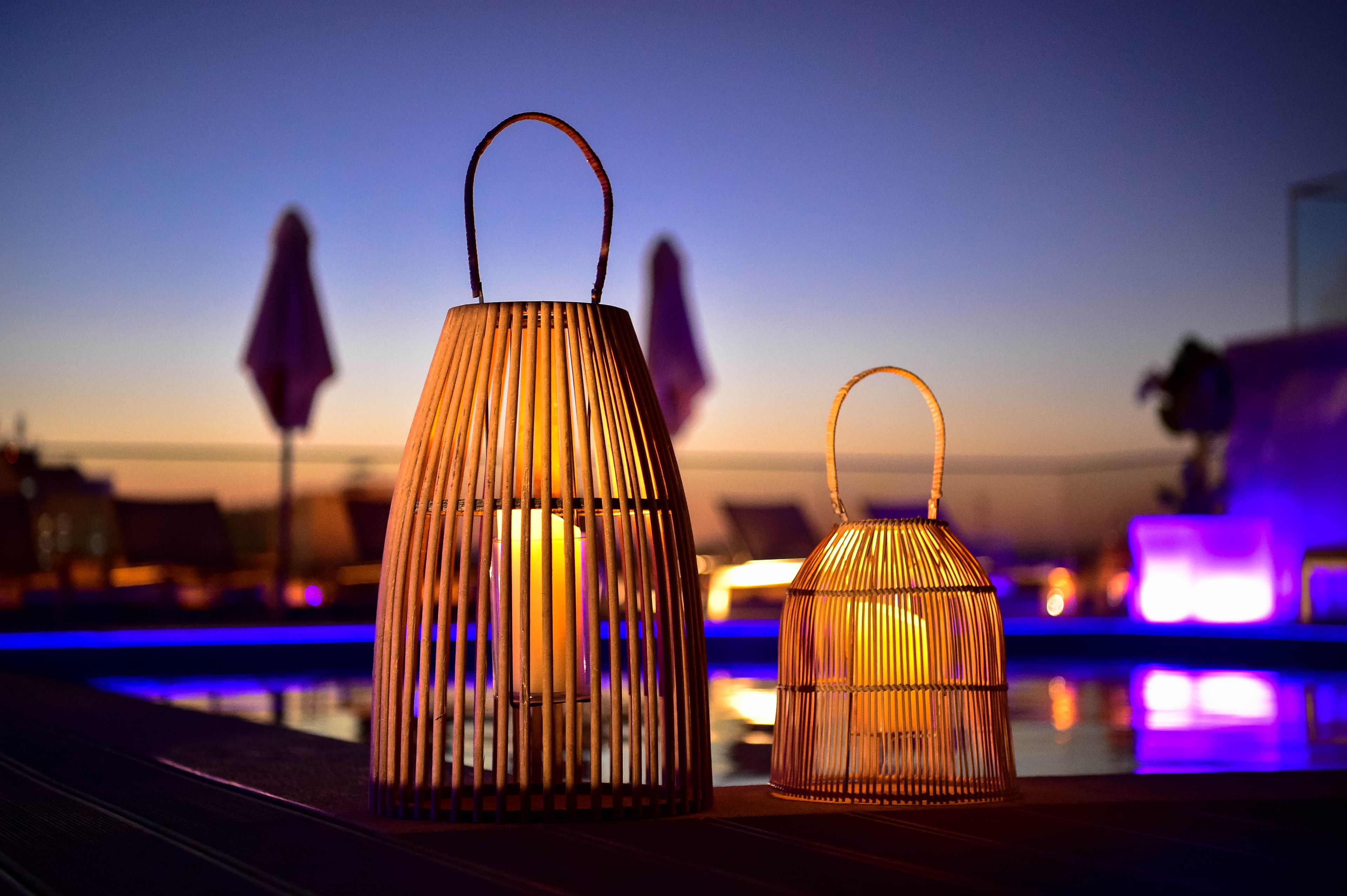 Deux lanternes en osier, dotées d’anses, sont posées sur un balcon. On aperçoit une piscine et les lumières de la ville en arrière-plan.