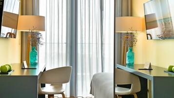 Habitación de hotel con escritorio, dos sillas, una cama, un ventanal grande con cortinas traslúcidas y un televisor montado en la pared.