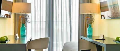 A hotel room with a desk, two chairs, a bed, a large window with sheer curtains, and a wall-mounted TV.