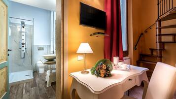 Ein modernes Badezimmer mit Glasdusche, Toilette, Waschbecken, einem Fernseher über dem Spiegel und einem Tisch mit Lampe und Blumenstrauß.