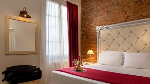 Hotelkamer met een bakstenen muur, een groot bed met witte lakens, een rode plaid, een nachtkastje met een lamp en een koffer op een stoel.
