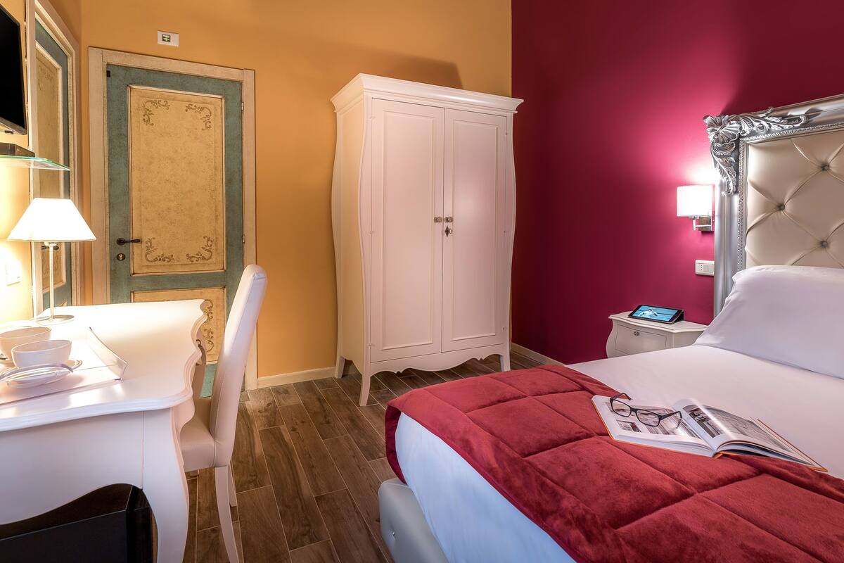 A hotel room with a bed, a desk with a lamp, a chair, a wardrobe, and a bedside table with a tablet.