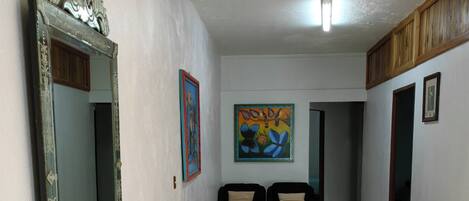 Un pasillo con suelo de baldosas, un sofá negro, un espejo grande y un cuadro colorido en la pared.