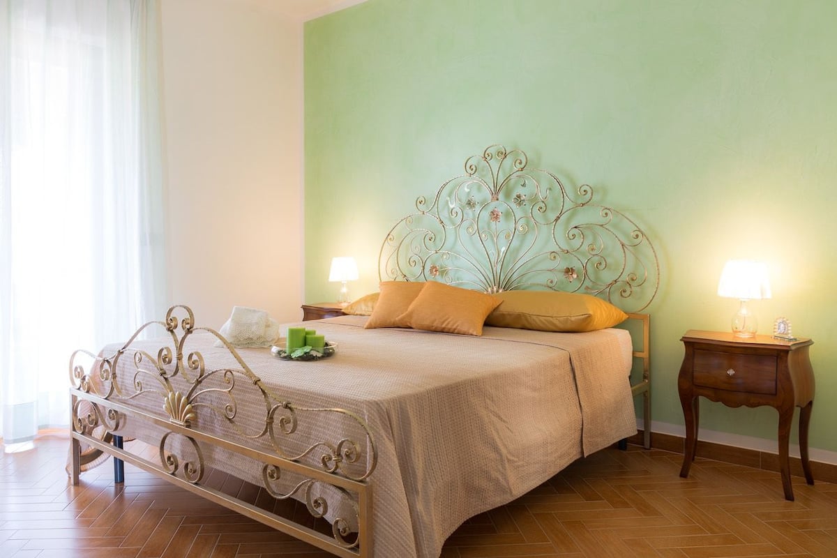 Una cama bien hecha con cabecera de metal, una mesita de noche con lámpara y una pared verde.