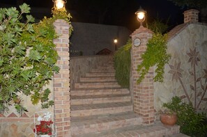 Un conjunto de escalones de piedra que conducen a la entrada de un edificio, flanqueados por pilares de ladrillo con luces, rodeado de vegetación.