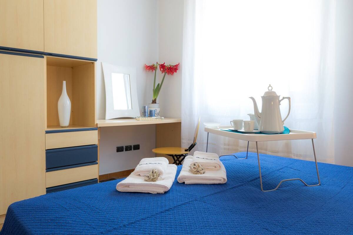 Habitación de hotel con cama, escritorio con cafetera, jarra con flores y ventana con cortinas.