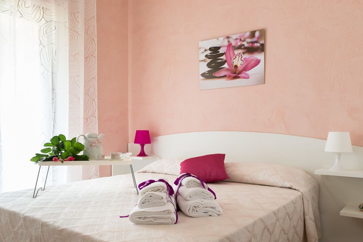 Una cama bien hecha con toallas enrolladas, una almohada rosa y un cuadro en la pared con una flor y piedras.