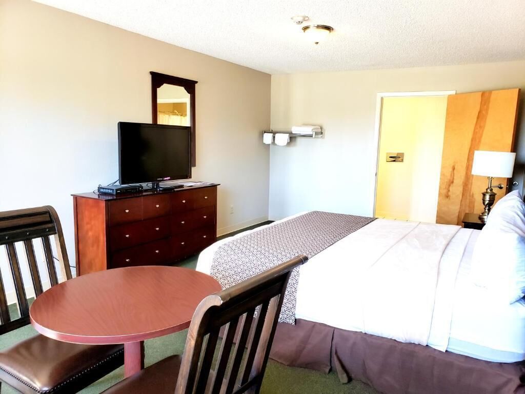 A hotel room with a bed, a desk with a television, a dresser, a chair, and a lamp.