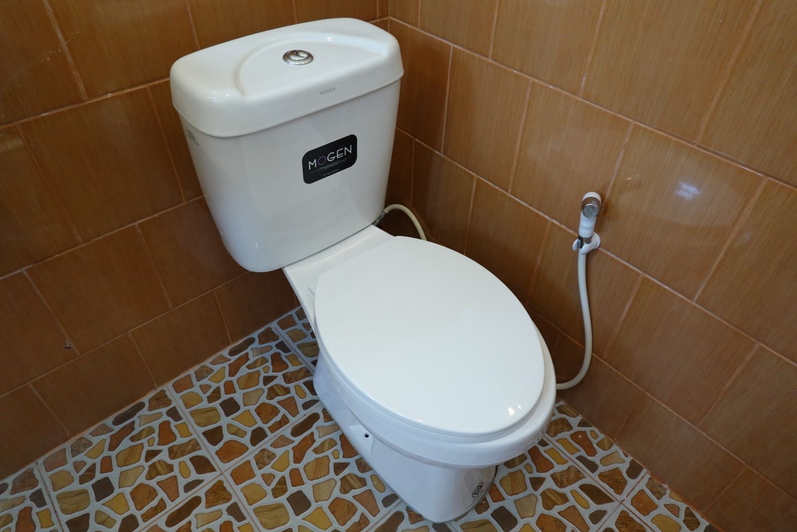 Toilet met een witte zitting en tank, voorzien van de tekst 'MOGEN', in een badkamer met bruine betegelde wanden en een mozaïek vloer.