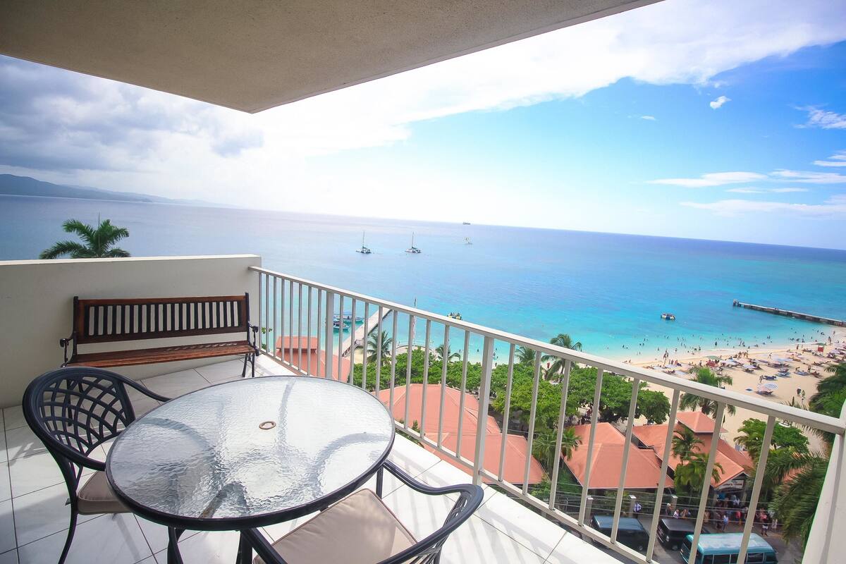 A balcony with a table and chairs, overlooking a beach with boats and a pier.