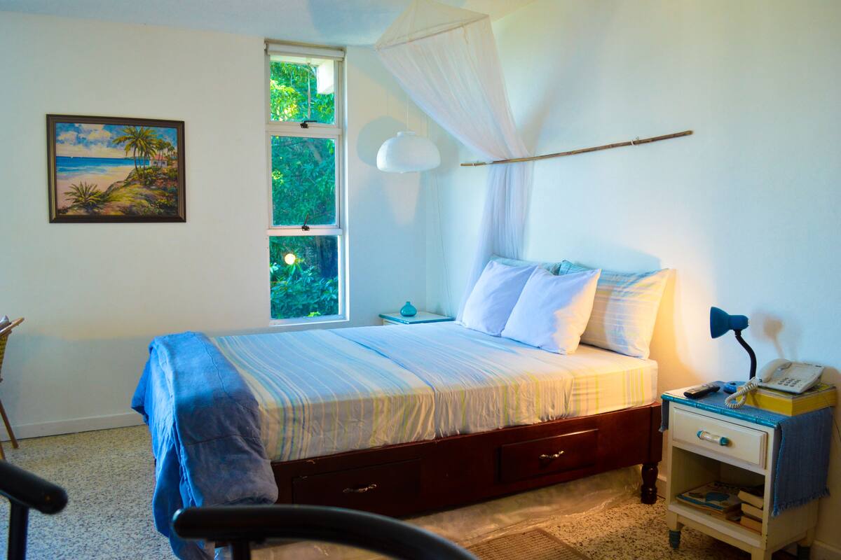 A bedroom with a bed, a nightstand, a chair, a window with a view of greenery, and a painting on the wall.