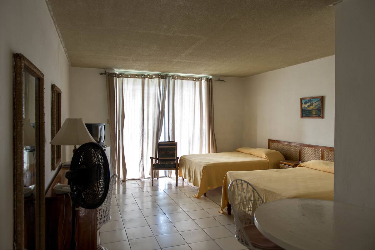 A hotel room with two beds, a chair, a table, a fan, and curtains.