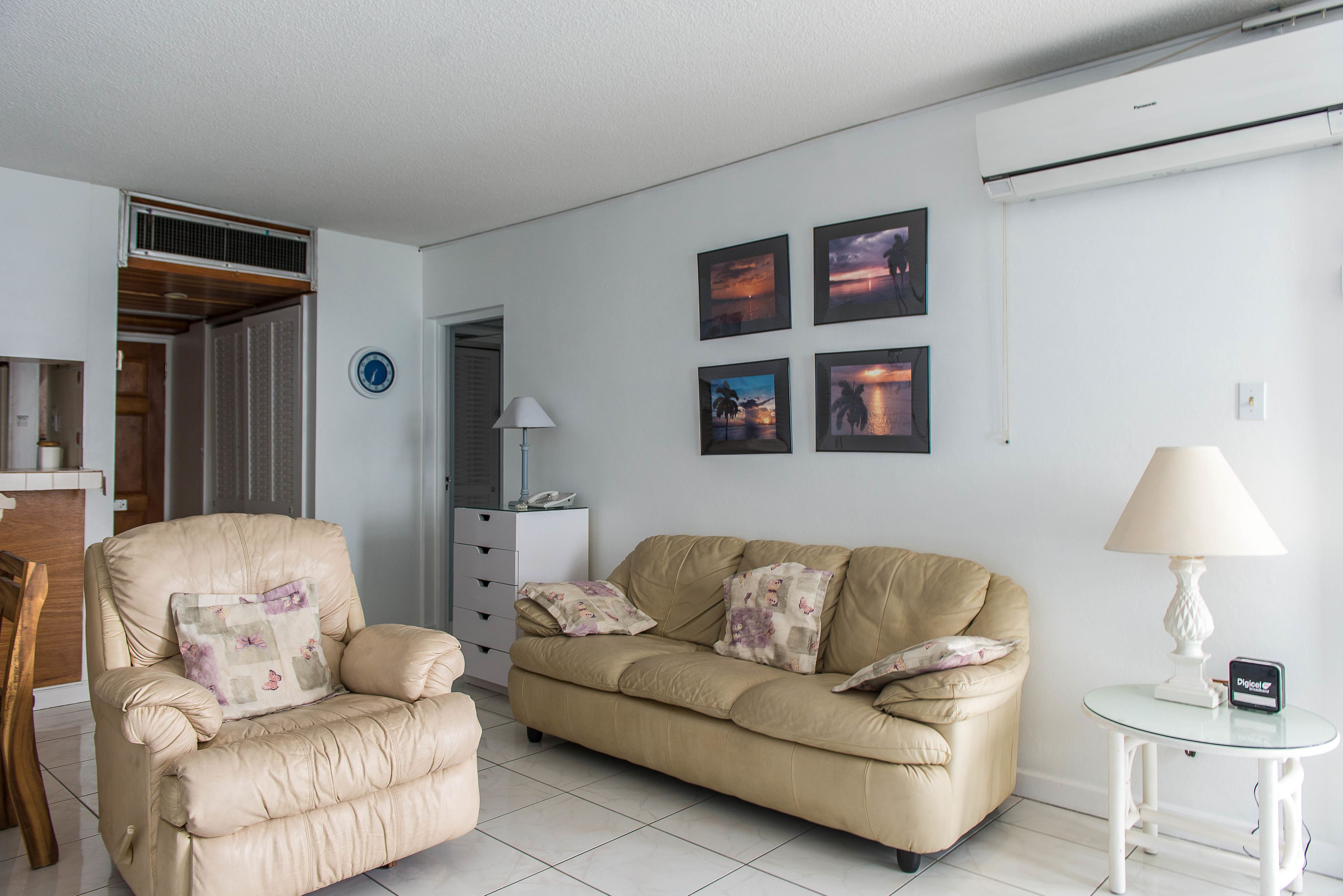 Un salon avec un canapé beige, un fauteuil assorti, une petite table avec une lampe et un climatiseur fixé au mur.