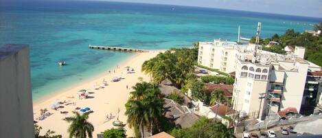A beach with clear turquoise waters, a pier extending into the sea, and a hotel with multiple floors and balconies overlooking the beach.
