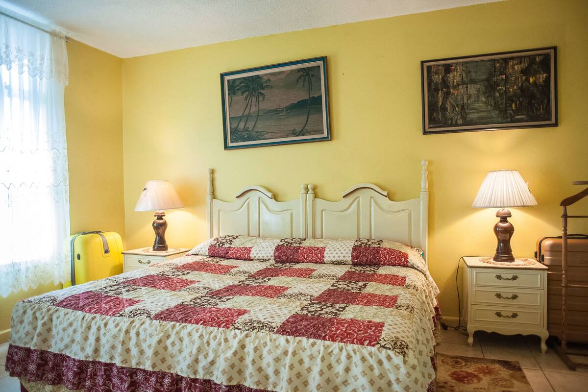 A bedroom with a bed, two bedside tables, a lamp on each table, a suitcase, and two framed pictures on the wall.