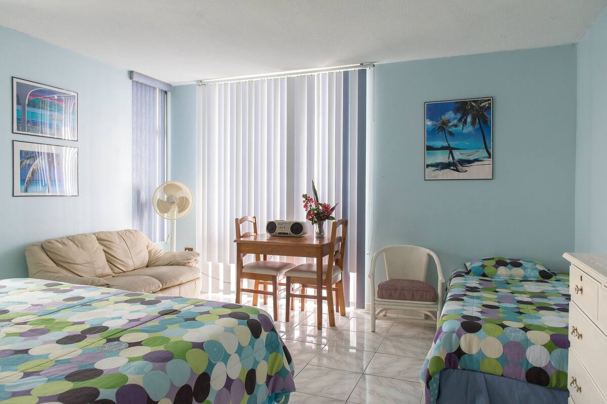 A hotel room with two beds, a dining table with chairs, a fan, and a beach-themed painting.