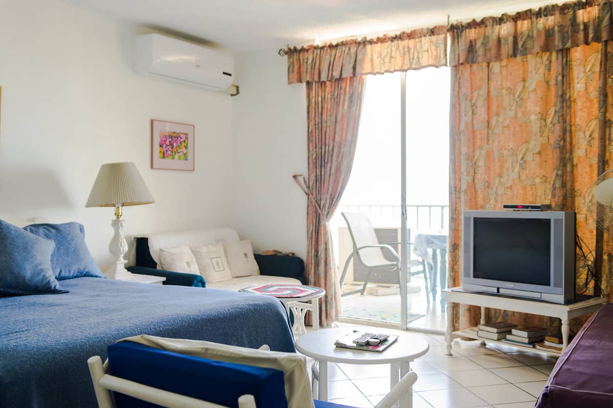 A hotel room with a bed, a small table, a TV on a stand, and a balcony with a chair.