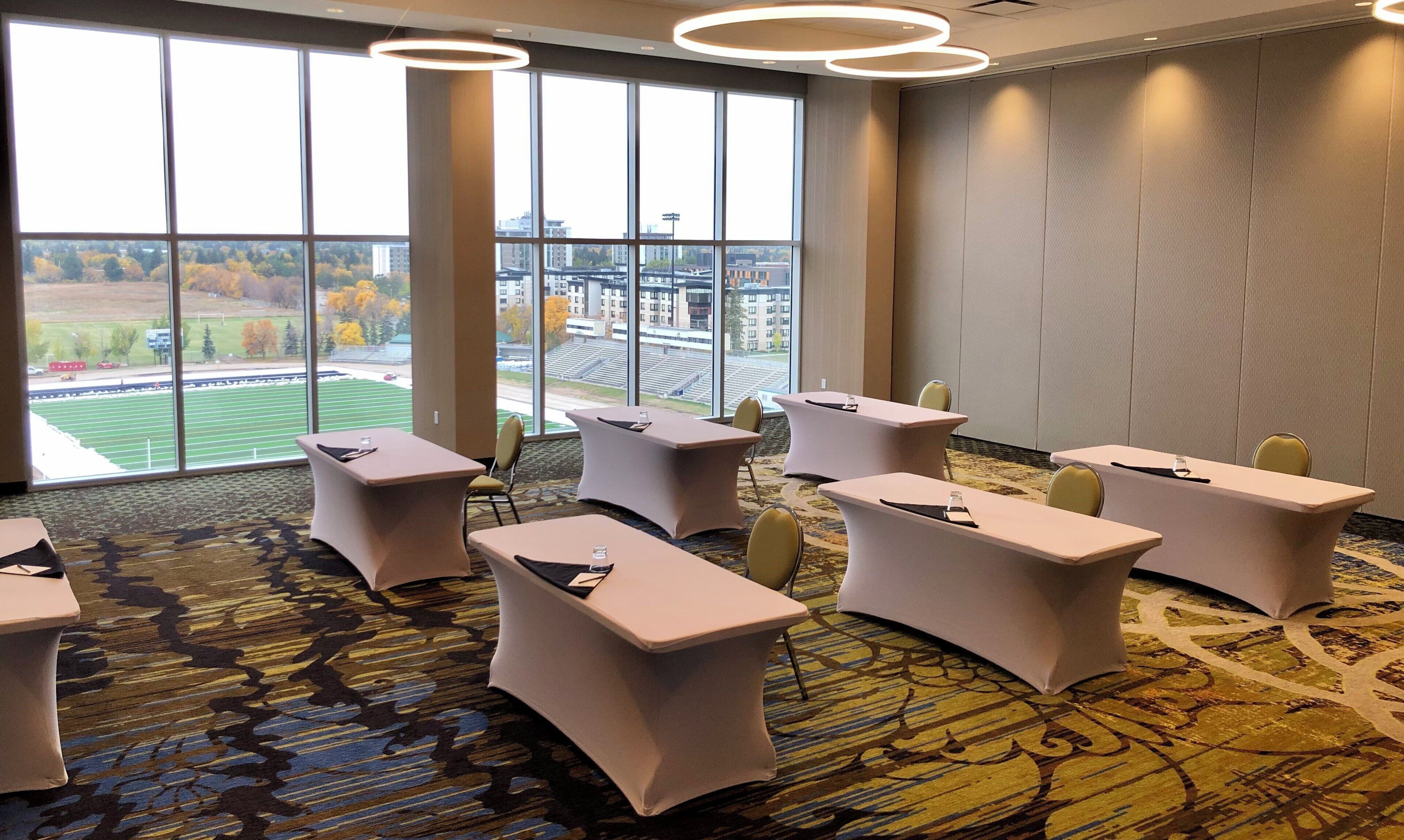 A conference room with tables and chairs, large windows with a view of a sports field, and a patterned carpet.
