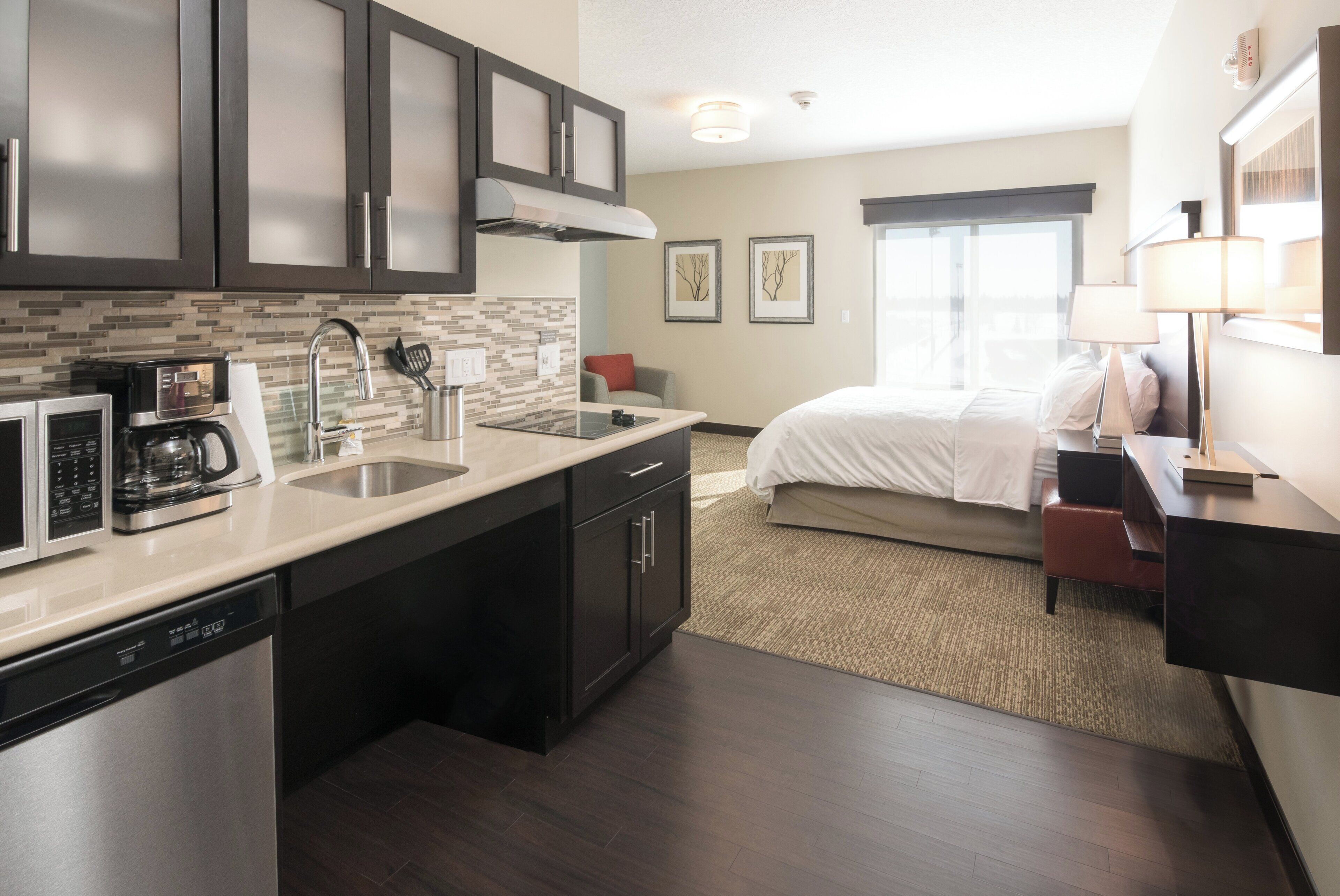 A modern hotel room with a kitchenette, a bed, a desk, and a window with a view.
