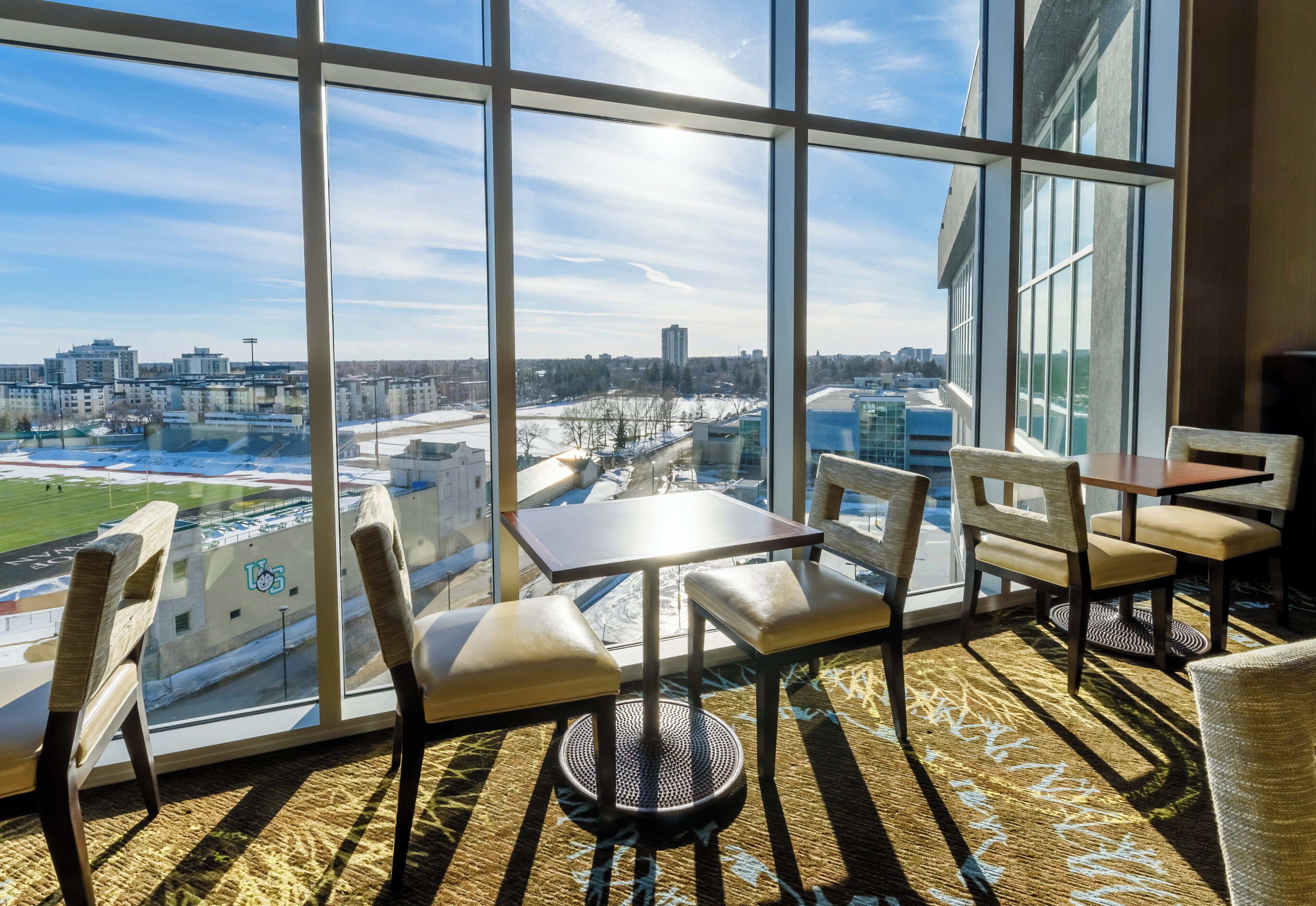 A modern interior with large windows offering a city view, a dining area with chairs and tables, and a patterned carpet.